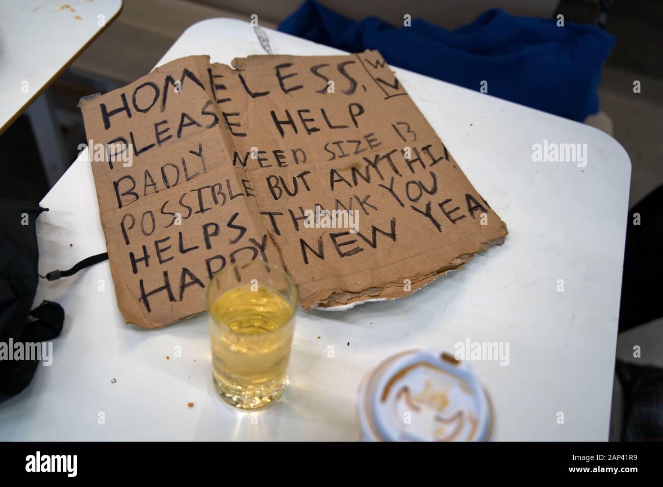 A homeless man`s sign sitting on a restaurant table beside refreshments and personal belongings. Stock Photo